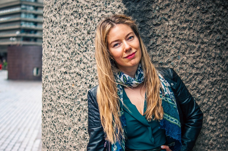 Researcher Alisa Kazarina standing outdoors, leaning against a textured wall and smiling at the camera.